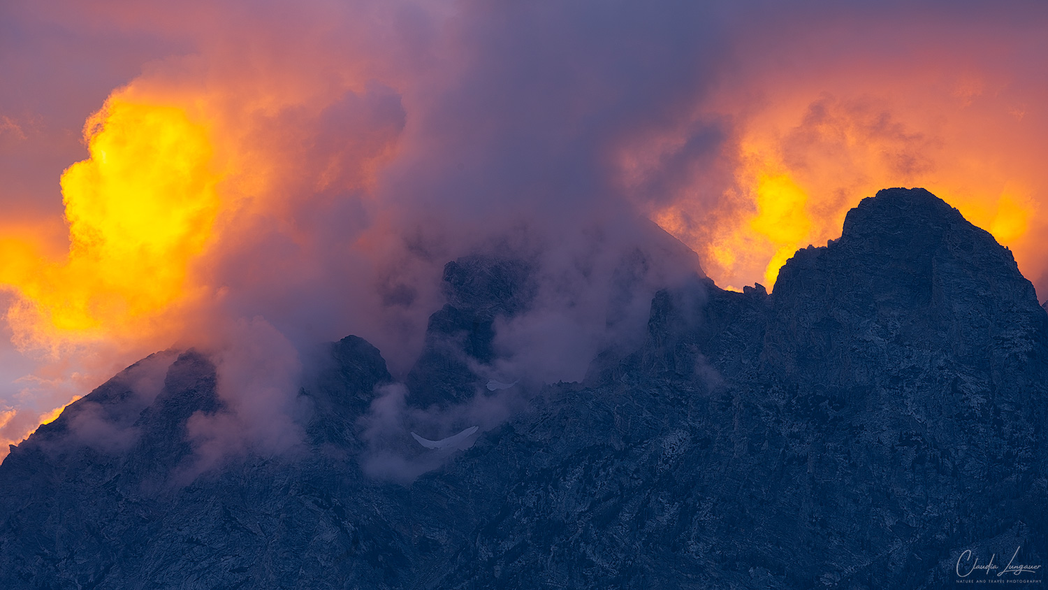 Fire in the Clouds Dramatic mountain peaks beneath swirling clouds illuminated by brilliant golden-orange sunset light, contrasting with the deep blue rocky slopes below.
