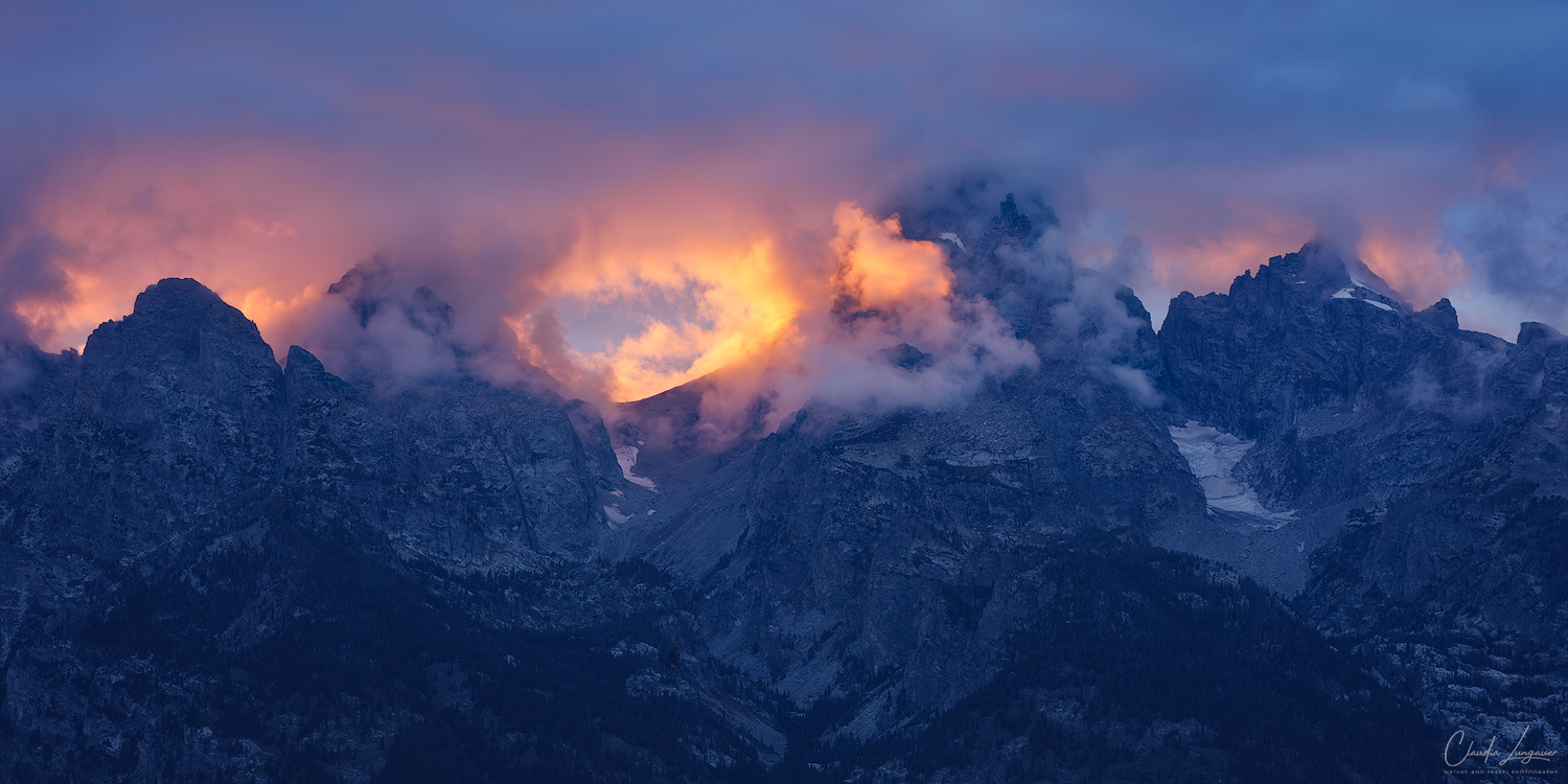 Where the Mountains Hold the Fire Panoramic sunset photograph of the Teton Range in Grand Teton National Park, Wyoming, with glowing orange and pink clouds swirling around rugged mountain peaks